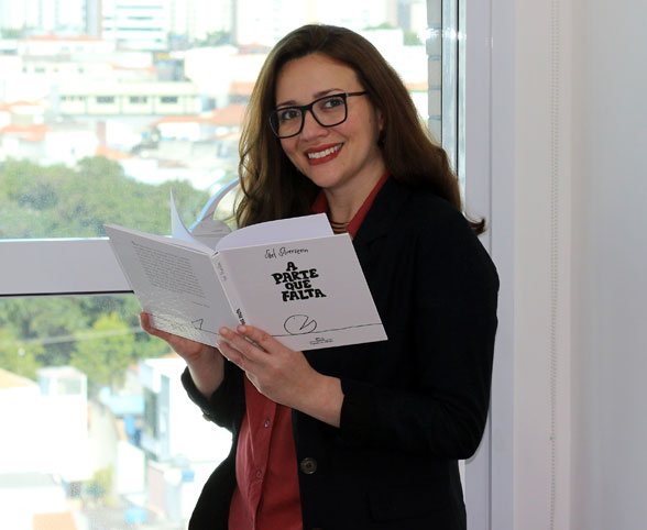 Fotografia da psicóloga Caroline Cunsolo em frente a uma janela de um consultório com o livro A Parte de Falta em suas mãos.